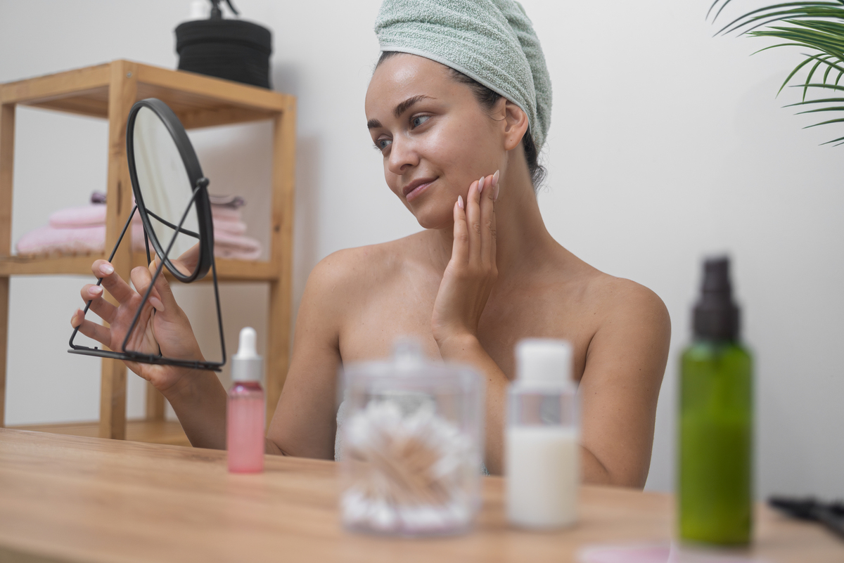 woman in towel applying skincare in a hand held mirror